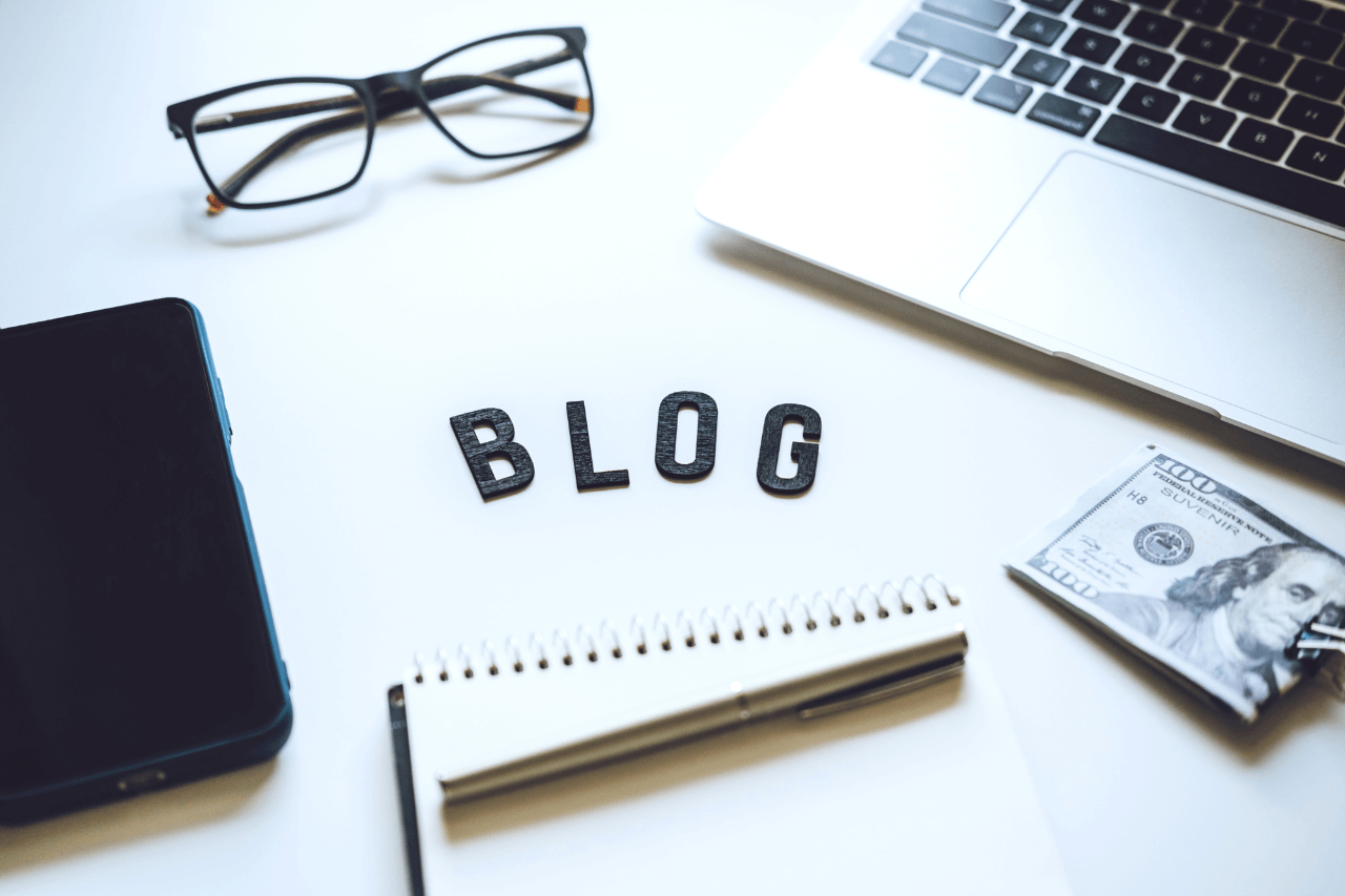 An organized desk with a laptop, glasses, phone, notebook, and $100, symbolizing the beginning of a blogging journey.
