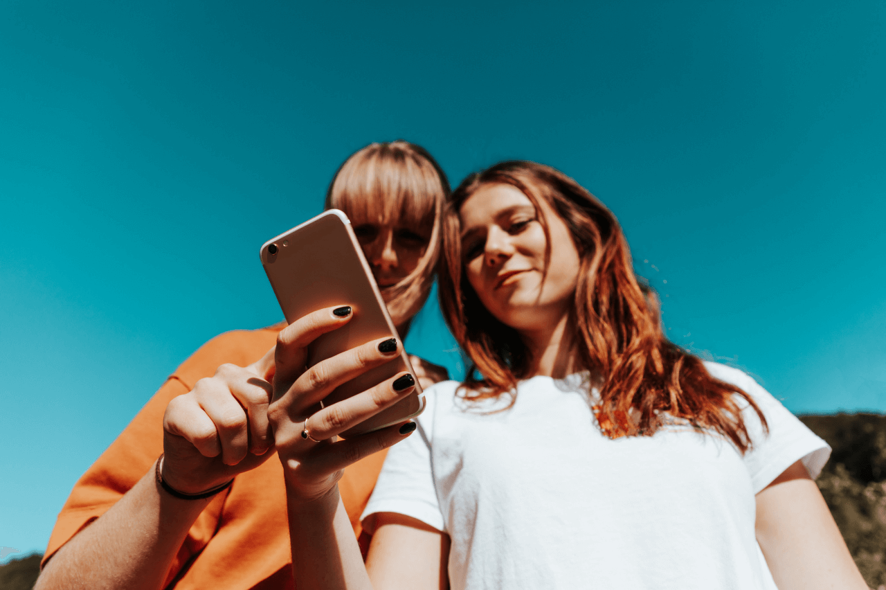 Two women standing side by side, engaged in conversation while using a cell phone together.